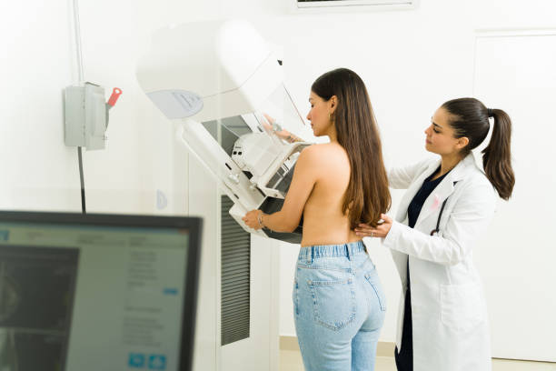 Medical professional guides a patient through a mammography exam with the imaging unit beside them, in a clinical setting.
