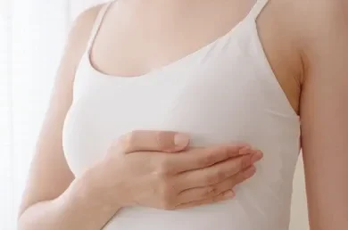 Close-up of a person in a white tank top placing a hand over their chest, as if performing a breast self-exam.