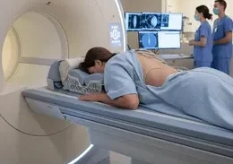 Patient lying on MRI table preparing for scan as a large MRI machine stands beside with medical staff and monitors in the background