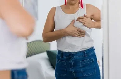Woman in a white sleeveless top and blue jeans adjusts her chest while looking at herself in a mirror in a bedroom.
